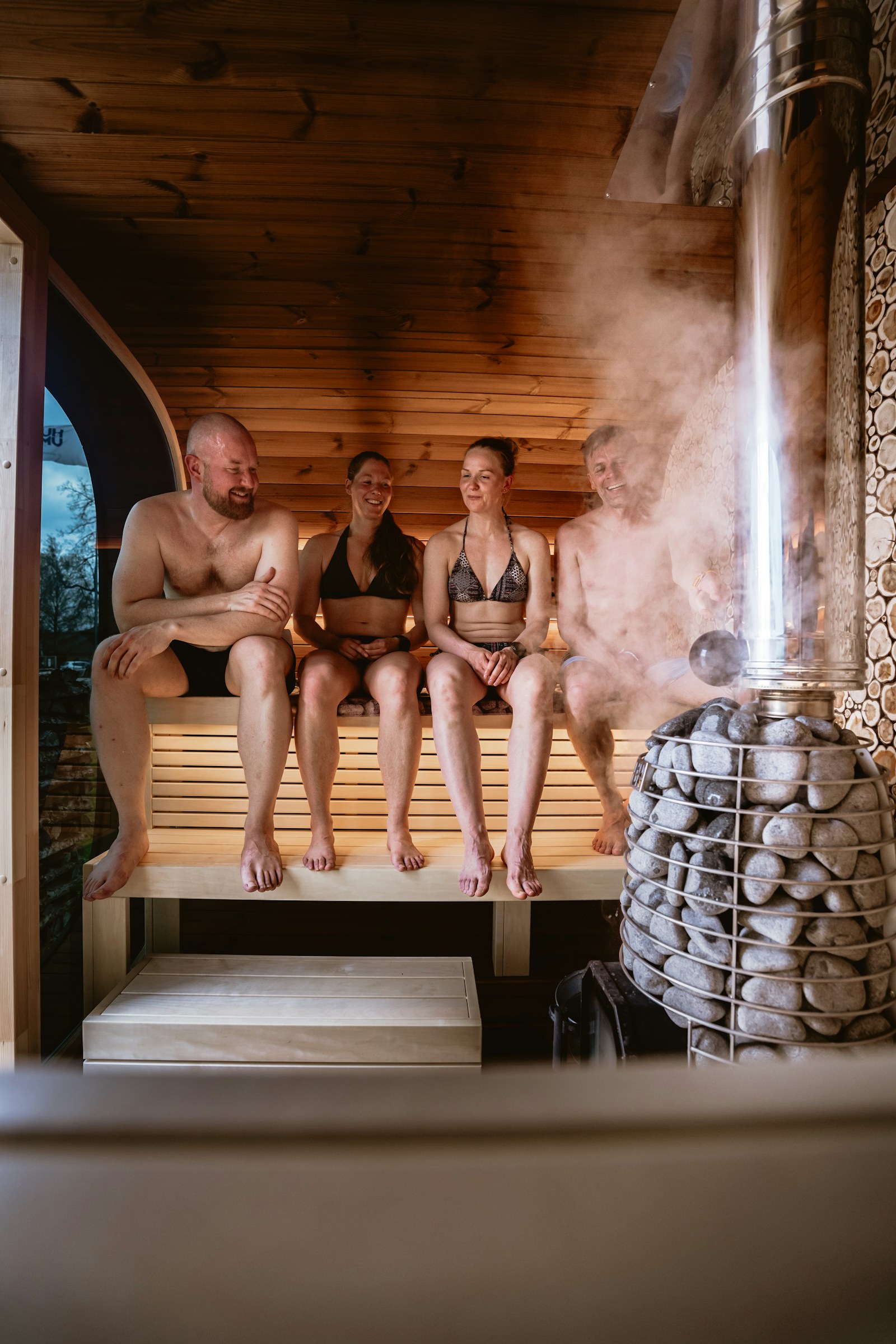Four people sitting on the benches of a wood-lined sauna as steam rises off the stove